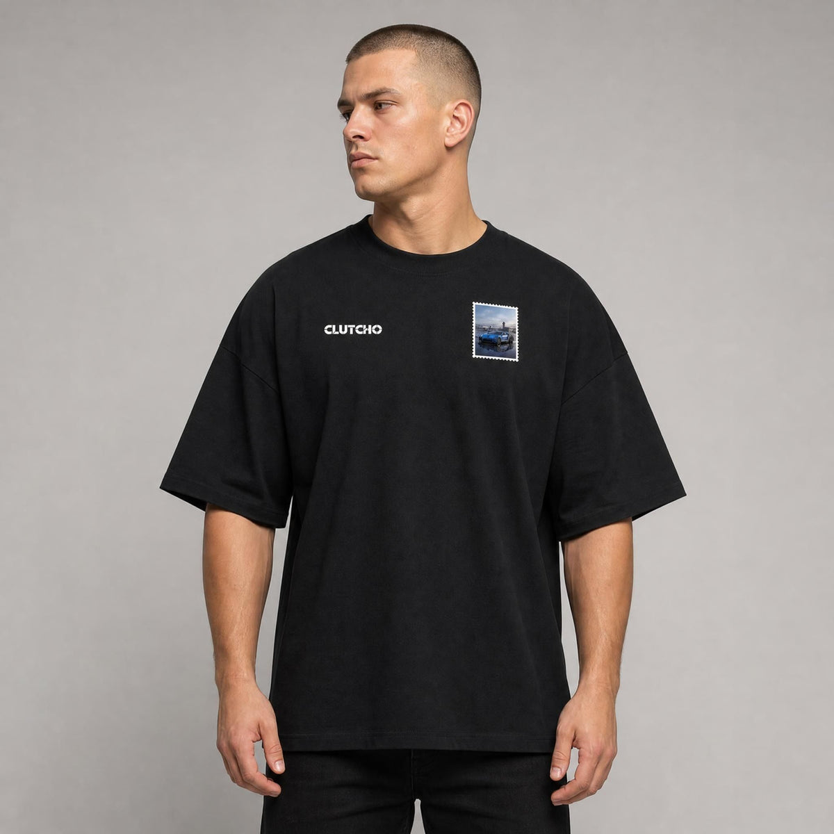 Schwarzes CLUTCHO Jetset Oversized T-Shirt Herren, Front-Briefmarke mit Hypercar auf dem Tarmac eines Privatflughafens.