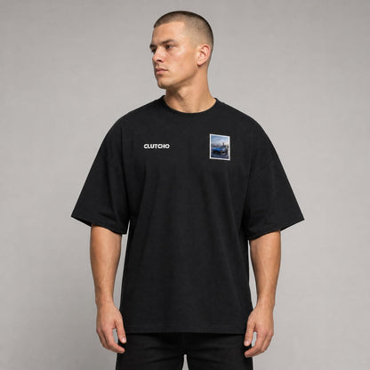 Schwarzes CLUTCHO Jetset Oversized T-Shirt Herren, Front-Briefmarke mit Hypercar auf dem Tarmac eines Privatflughafens.