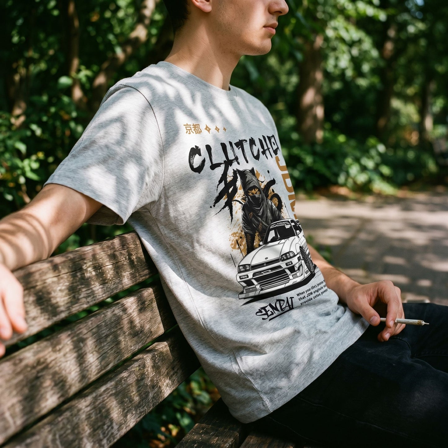 Profile view of model wearing CLUTCHO Senpai JDM T-Shirt on wooden bench in park with greenery.